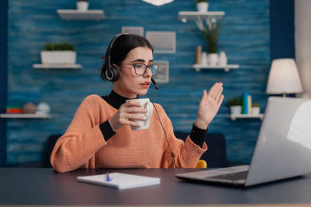 Student with glasses casually waving hello in laptop videocall. Person with headphones raising hand...