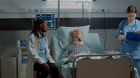 Multi ethnic medical staff checking on patient healthcare in hospital ward bed at intensive care. African american doctor helping old man with cervical neck collar and oxygen tubeの写真素材