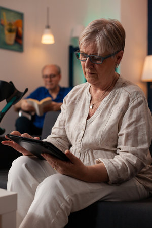 Aged person with crutches looking at digital tablet on couch at home. Elderly woman using modern device while senior man sitting in wheelchair reading book in living room togetherの写真素材