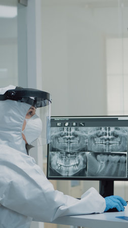Professional dentistry staff with ppe suit looking at teeth x ray in dental cabinet. Assistant examining teeth radiography on computer monitor while doctor preparing for consultationの写真素材