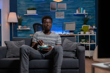 African american guy resting on sofa in living room holding popcorn bowl while watching entertainment movie on television. Young man enjoying spending free time alone in living roomの写真素材