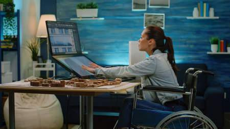 Disabled architect analyzing building model on touch screen monitor for construction layout. Engineer with handicap working on blueprint plan while sitting in wheelchair. Architectural equipmentの写真素材