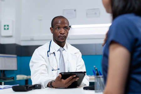 African american pediatrician doctor typing sickness symptoms on tablet computer discussing healthcare treatment with mother during clinical examination. Therapist man working in hospital officeの写真素材