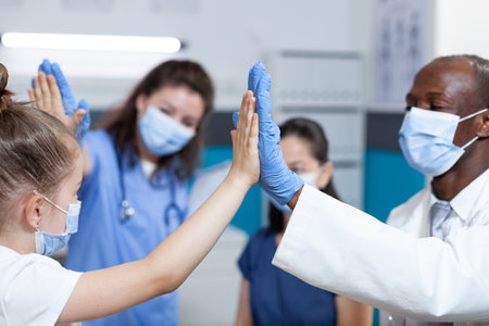 Young girl patient with protection face mask against covid19 giving high five to medical team during clinical consultation in hospital office. Pediatric doctor discussing healthcare treatmentの写真素材