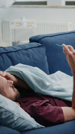 Man with headache holding thermometer for fever measurement and resting on couch. Sick person using medical device to check temperature for treatment against coronavirus illnessの写真素材