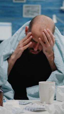 Close up of sick adult with headache rubbing temples in blanket. Man with migraine and flu symptoms trying to cure pain while having medicaments, pills, tea and thermometer on tableの写真素材