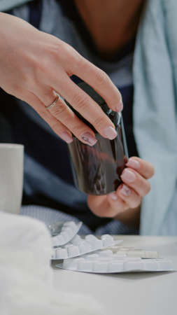 Close up of woman reading labels of tablets and jars with pills and capsules on table with medicaments, tissues and thermometer. Sick person looking for treatment to cure virus symptomsの写真素材