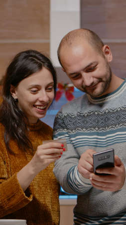 Festive couple buying presents with credit card on smartphone, doing online shopping for christmas eve celebration. People using device paying for gifts in seasonal decorated kitchenの写真素材