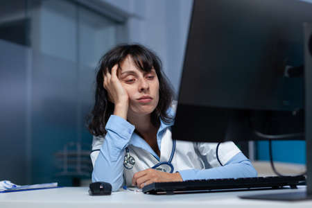 Doctor with closed eyes at medical office after hours, working late at night. Exhausted woman looking at computer screen, having head in hand doing overtime work for healthcare jobの写真素材