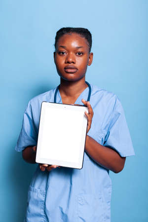Portrait of african american practitioner nurse in medical uniform holding tablet computer with white screen working at healthcare treatment in studio with blue baclground. Medicine conceptの写真素材