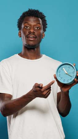 Casual person pointing at time on wall clock for hour and minutes. Portrait of young man looking at watch and explaining countdown and delay for work. Punctual adult setting alarmの写真素材