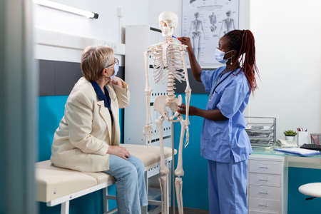 Physician nurse with protective face mask explaining anatomy body structure using medical skeleton discussing back pain with retired old woman during appointment in hospital office. Medicine conceptの写真素材