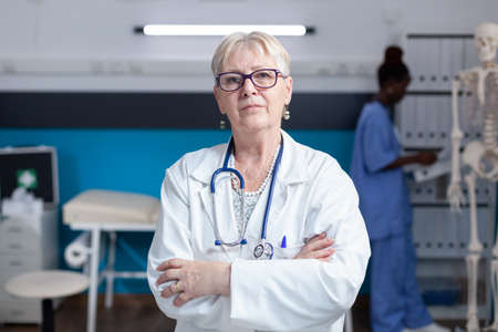Portrait of senior medic standing with arms crossed in healthcare cabinet, having stethoscope. Close up of specialist ready to practice medicine and medical expertise. Confident woman doctorの写真素材