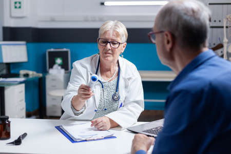 Medic looking at thermometer after checking fever measurement for old patient. Woman doctor...
