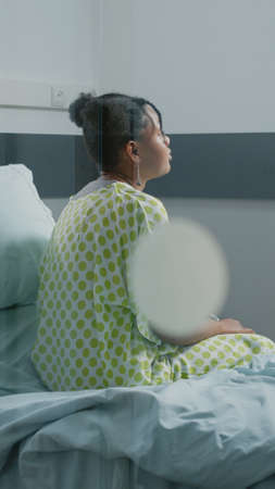 Portrait of young adult with sickness sitting in hospital ward bed to receive medical care and treatment. Patient waiting on medic to help with pain and disease at healthcare clinic.の写真素材