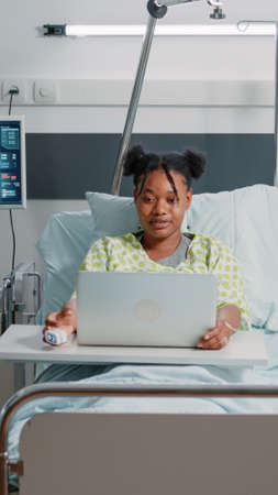 African american woman waving at video call on laptop in hospital ward. Young adult using online conference on device to communicate with friends and family. Remote conversation on gadgetの写真素材