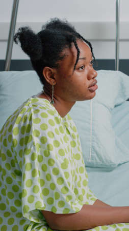 Portrait of african american patient sitting in hospital ward bed, waiting on doctor to give medical support and treatment to cure disease. Young woman with IV drip bag feeling sickの写真素材