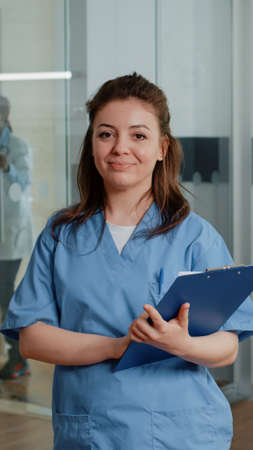 Portrait of woman working as nurse holding checkup papers while standing in hospital ward. Medical assistant with documents looking at camera and smiling, giving help to specialist.の写真素材