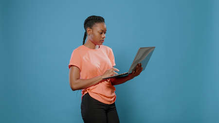 Young woman holding modern laptop in front of camera. Person using wireless computer and technology while standing over blue background. Adult holding gadget and looking at displayの写真素材