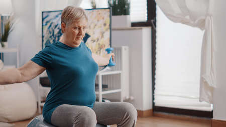 Close up of pensioner using dumbbells on fitness toning ball, training and stretching arms muscles at home. Aged woman doing physical exercise, practicing sport with equipment.の写真素材