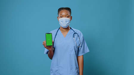 Medical assistant vertically holding smartphone with green screen and looking at camera. Nurse with face mask showing isolated background and mockup template with chroma key on display.の写真素材