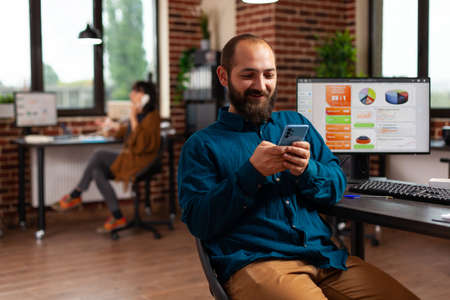 Executive manager sitting at desk table holding modern phone reading email messaging with remote friends telling good news while working at management project. Businessman searching business informationの写真素材
