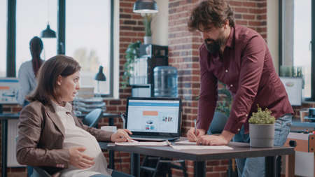 Pregnant woman doing teamwork with colleague for business project. Team of business people working together on startup development, using papers and laptop to design marketing strategy.の写真素材