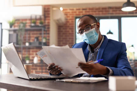 African american man with face mask analyzing documents to plan marketing strategy. Company employee working with laptop and doing paperwork for project during coronavirus pandemic.の写真素材