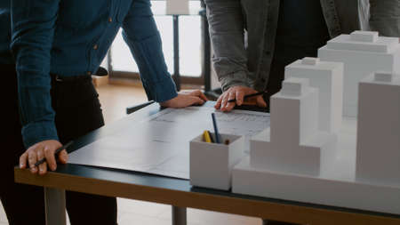 Close up of colleagues working on urban design with blueprints and building model. Team of people doing teamwork on construction layout, designing architectural development project.の写真素材