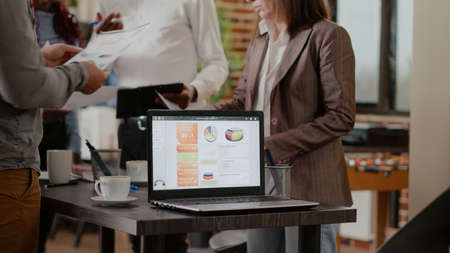 Close up of laptop with business charts on desk in startup office. Colleagues working together to...