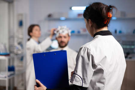 Doctor woman holding clipboard with neuroscience information on it while neurologist adjusting eeg headset monitoring nervous system of man patient. Physician engineer analyzing brain activityの写真素材