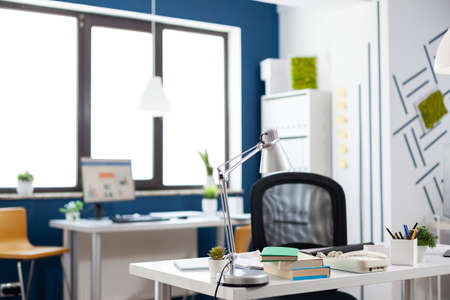 Lamp on a white desk in an empty modern business office. Startup workspace furniture with blue and white wall. Table in commercial workplace with big window. Nobody in indoors work location.の写真素材