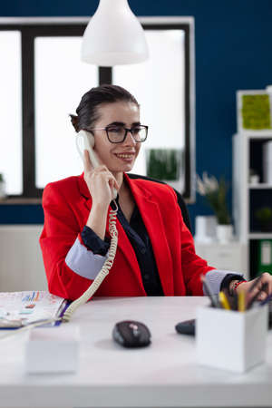 Smiling entrepreneur with glasses in phonecall sitting at startup desk. Businesswoman in red jacket listening to employee at the phone. Small business owner in casual conversation.の写真素材