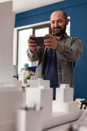 Professional architect holding smartphone smiling in videocall conference presenting final maquette to client. Bearded architectural engineer photographing table with white foam residential model.の写真素材