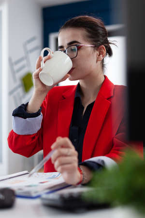 Manager in red jacket at startup desk drinking from white cup. Employee with glasses having coffe or tea break at the office. Businesswoman taking a pause from reading chart on clipboard.の写真素材