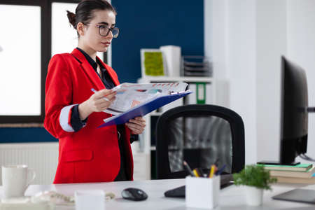 Businesswoman standing up next to desk holding clipboard with chart. Focused entrepreneur with glasses and red jacket in startup office looking at business results on computer screen.の写真素材
