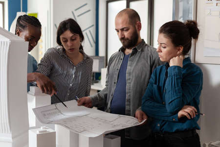 Team of architects analyzing blueprints for building project standing in front of table with real...