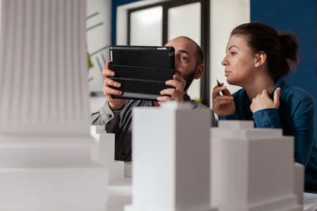 Project engineers working together looking at tablet with construction plans sitting at desk in modern architectural office. Two architects doing teamwork next to table with buildings maquette.の写真素材
