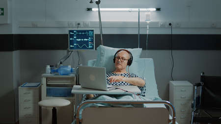 Sick old man sitting in hospital ward using laptop and headphones listening to music. Patient with disease relaxing while taking medical treatment from IV drip bag and nasal oxygen tubeの写真素材