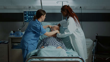 Patient going unconscious in hospital ward after surgery at medical facility. Nurse and african american doctor helping sick old man at reanimation room with oxygen tube and IV drip bagの写真素材