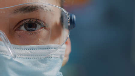 Scientist showing one eye on camera in laboratory, wearing safety glasses and face mask. Woman biologist looking at camera with face of face, working in research lab. Close upの写真素材