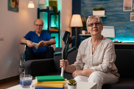 Portrait of aged woman sitting on couch and looking at camera while senior man in wheelchair reading...