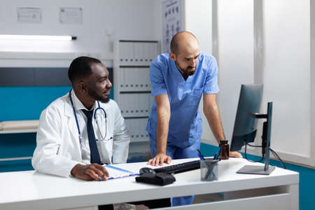 Team of medical therapists monitoring sickness symptoms expertise on computer discussing healthcare treatment. African american physician doctor working at medicine prescription in hospital officeの写真素材