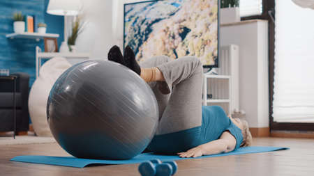Old woman stretching legs muscles on fitness toning ball to exercise on yoga mat. Retired person training with sport equipment and doing physical activity for wellness at home. Senior adultの写真素材