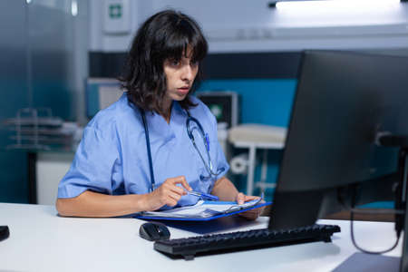 Medical assistant analyzing documents and files on monitor at night. Nurse looking at computer and working for healthcare appointment with checkup papers, doing overtime work at desk.の写真素材
