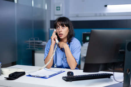Medical assistant talking on telephone to patient for appointment, working late. Woman nurse using...