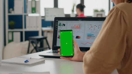 Close up of woman vertically holding smartphone with green screen in business office. Person looking at mobile phone with isolated mock up background and chroma key template on display.の写真素材