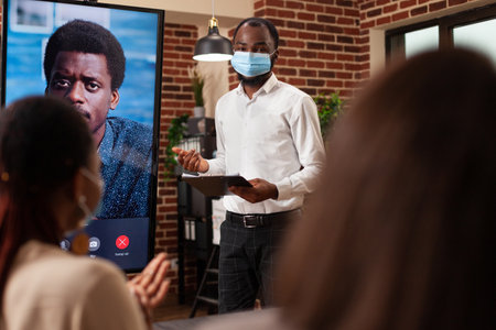 African american leader with medical face mask against covid19 explaining management strategy to businessteam in startup company office. Online videocall conference meeting on monitor screenの写真素材