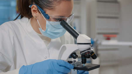 Biologist with face mask using microscope in science laboratory, analyzing dna sample and substance...