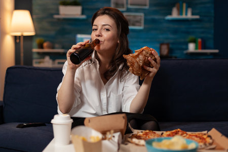 Woman in white shirt drinking beer from bottle while holding a tasty takeaway burger looking at television movie. Person sitting on couch eating delivery hamburger in front of table with fast food.の写真素材